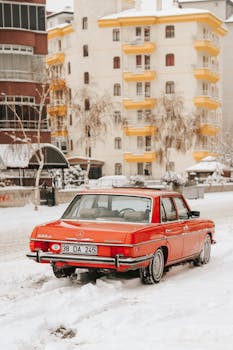 Classic red Mercedes parked on a snowy city street with residential buildings.