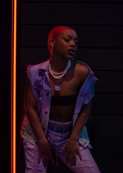 Stylish black woman with pink hair and pearls posing against a dark urban backdrop in Des Moines.