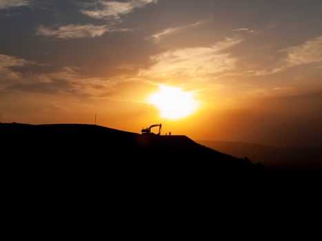 Silhouette of construction excavator on a hill at sunrise, creating a dramatic backlit scene.