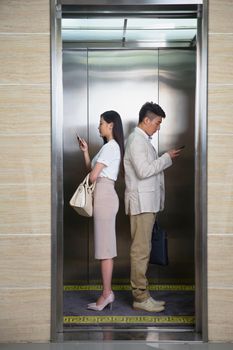 Two business professionals stand back to back in an elevator, both using phones.
