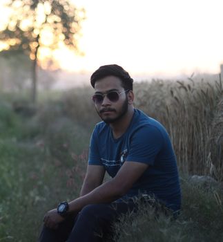 A young man in sunglasses sitting in a field, enjoying a calm sunset.