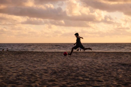 A child playing soccer on a sandy beach in Oaxaca, Mexico at sunset with dramatic clouds in the sky.