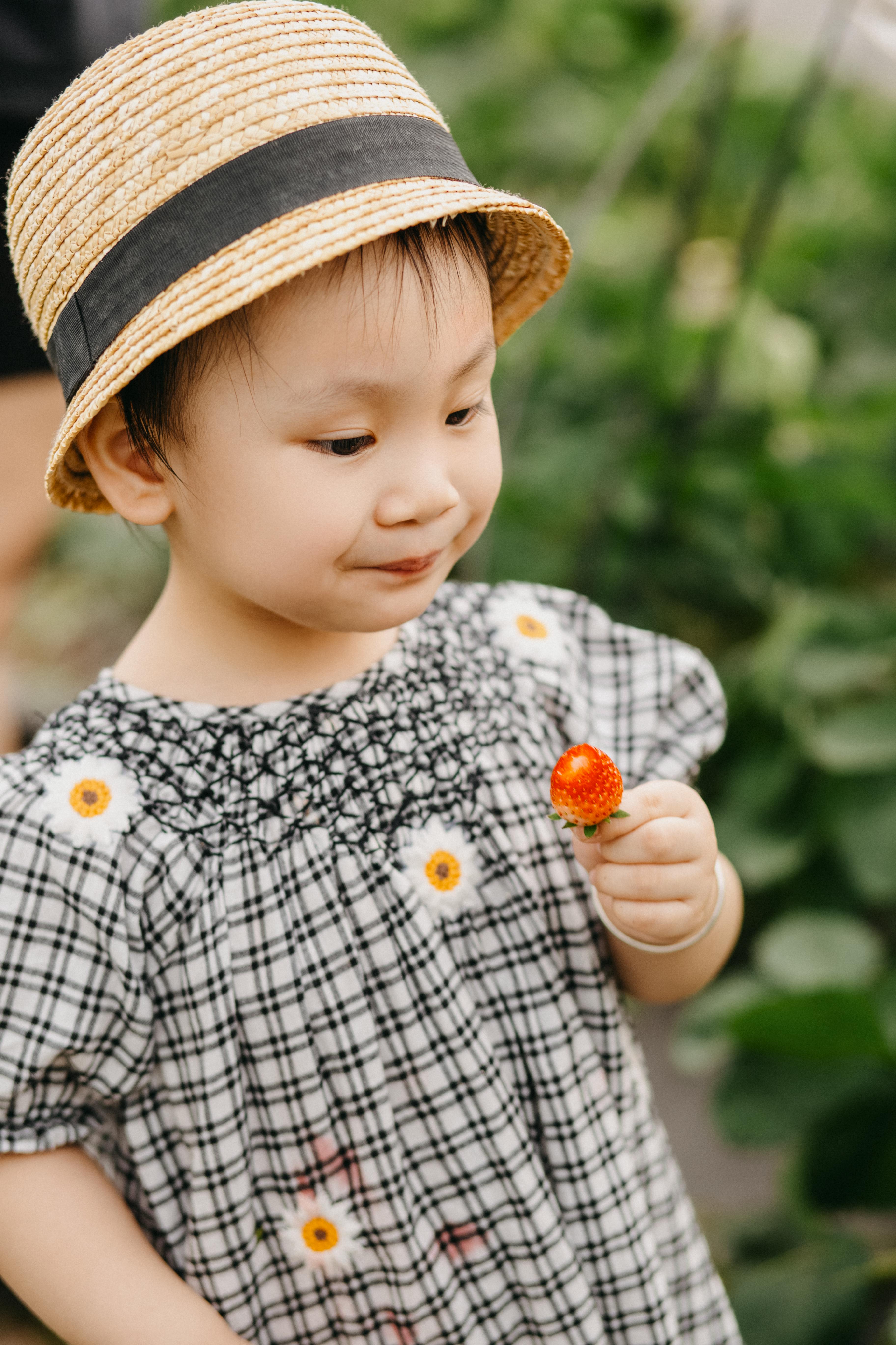 A charming young girl in a straw hat holds a strawberry in a garden setting.