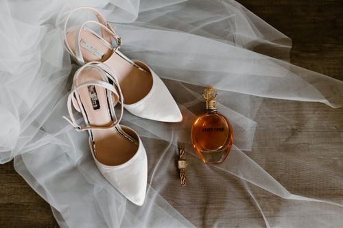 Free Chic wedding essentials displayed on sheer fabric include white pointed shoes, a stylish watch, and designer perfume. Stock Photo
