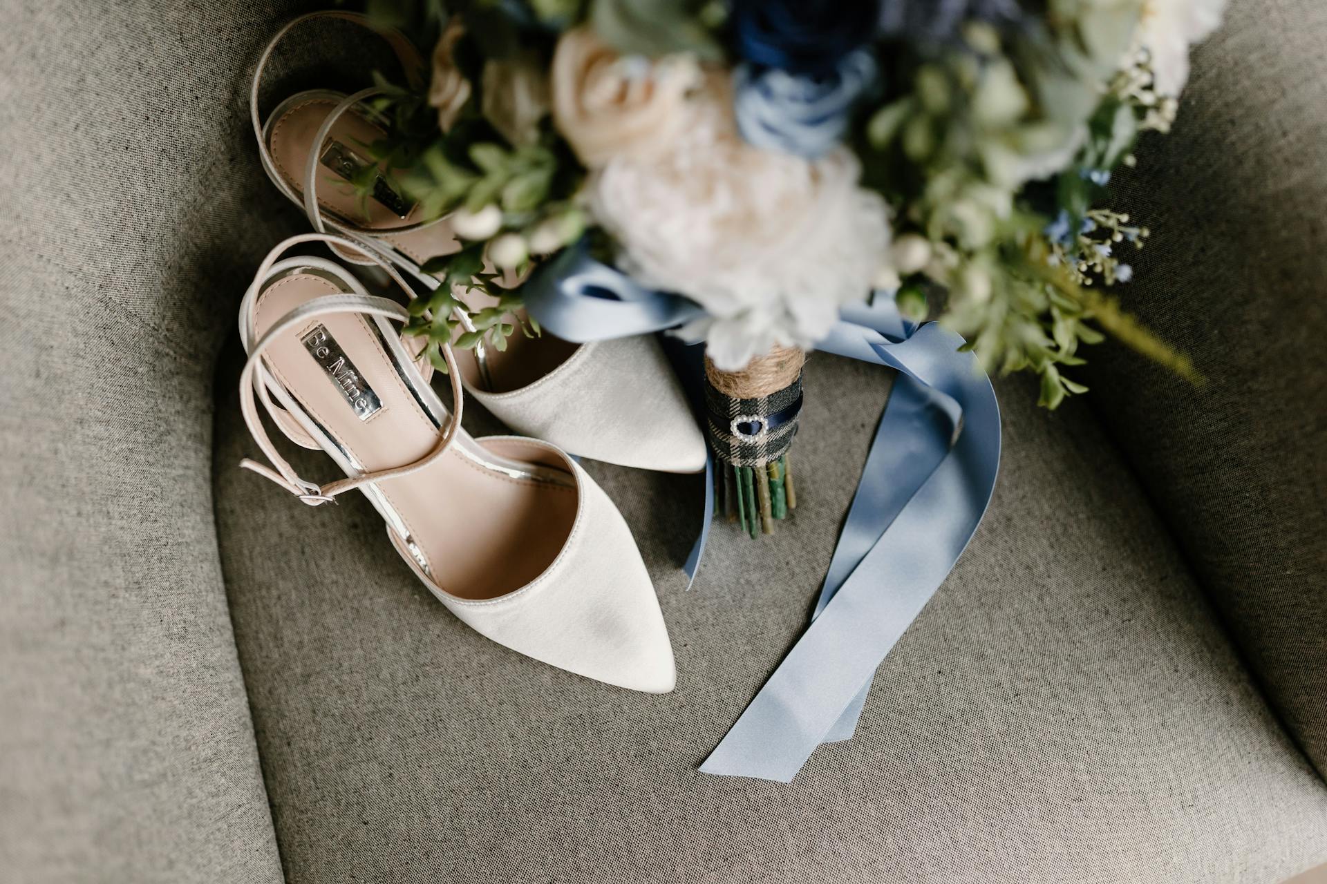 A top view of elegant white bridal shoes and a bouquet with a blue ribbon on a gray sofa.