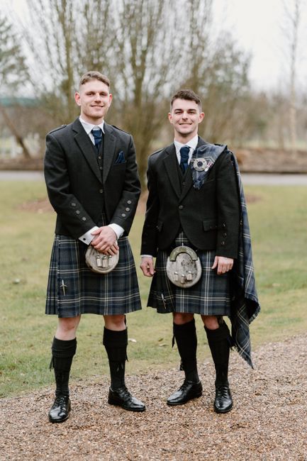 Two men in traditional Scottish kilts smiling and posing outdoors on a cool day.