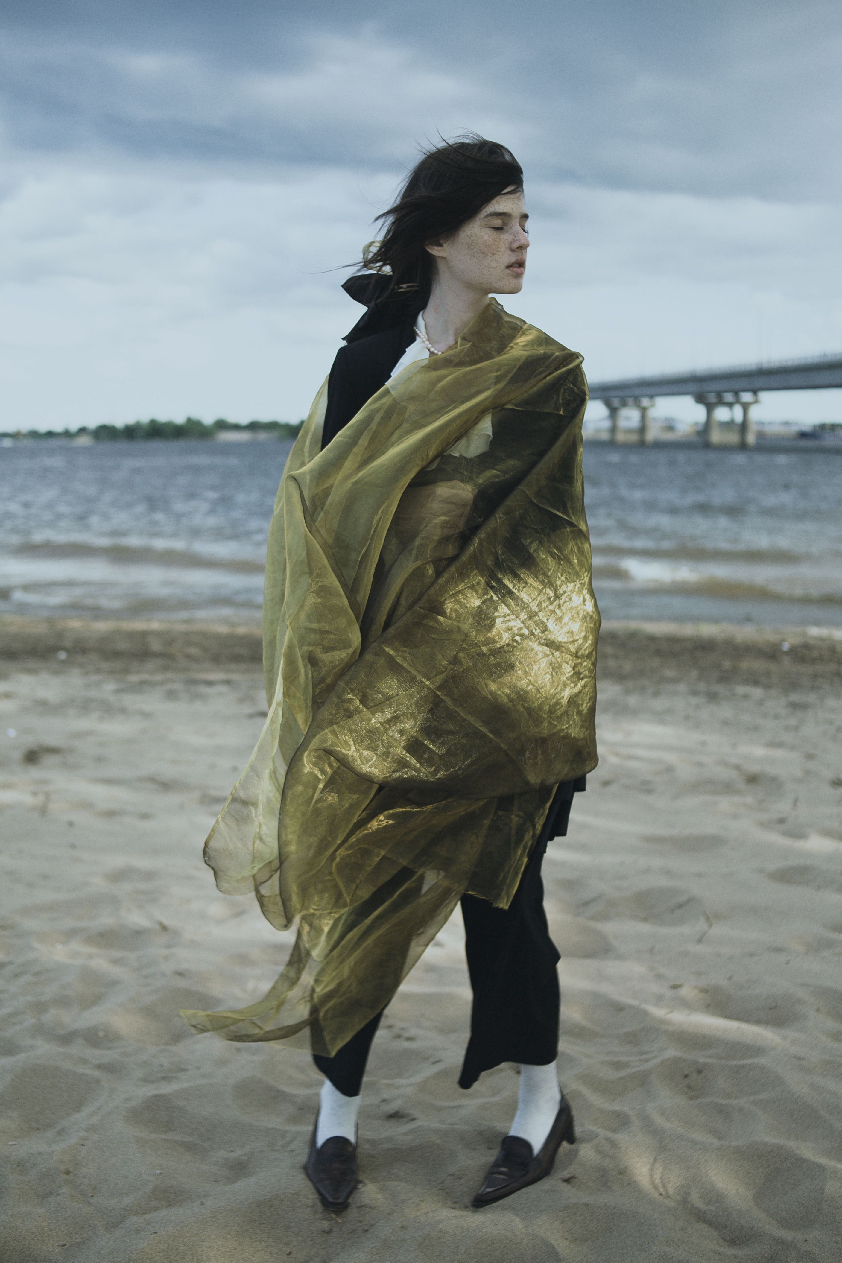 A woman stands on a beach wrapped in flowing green fabric with a bridge in the background.