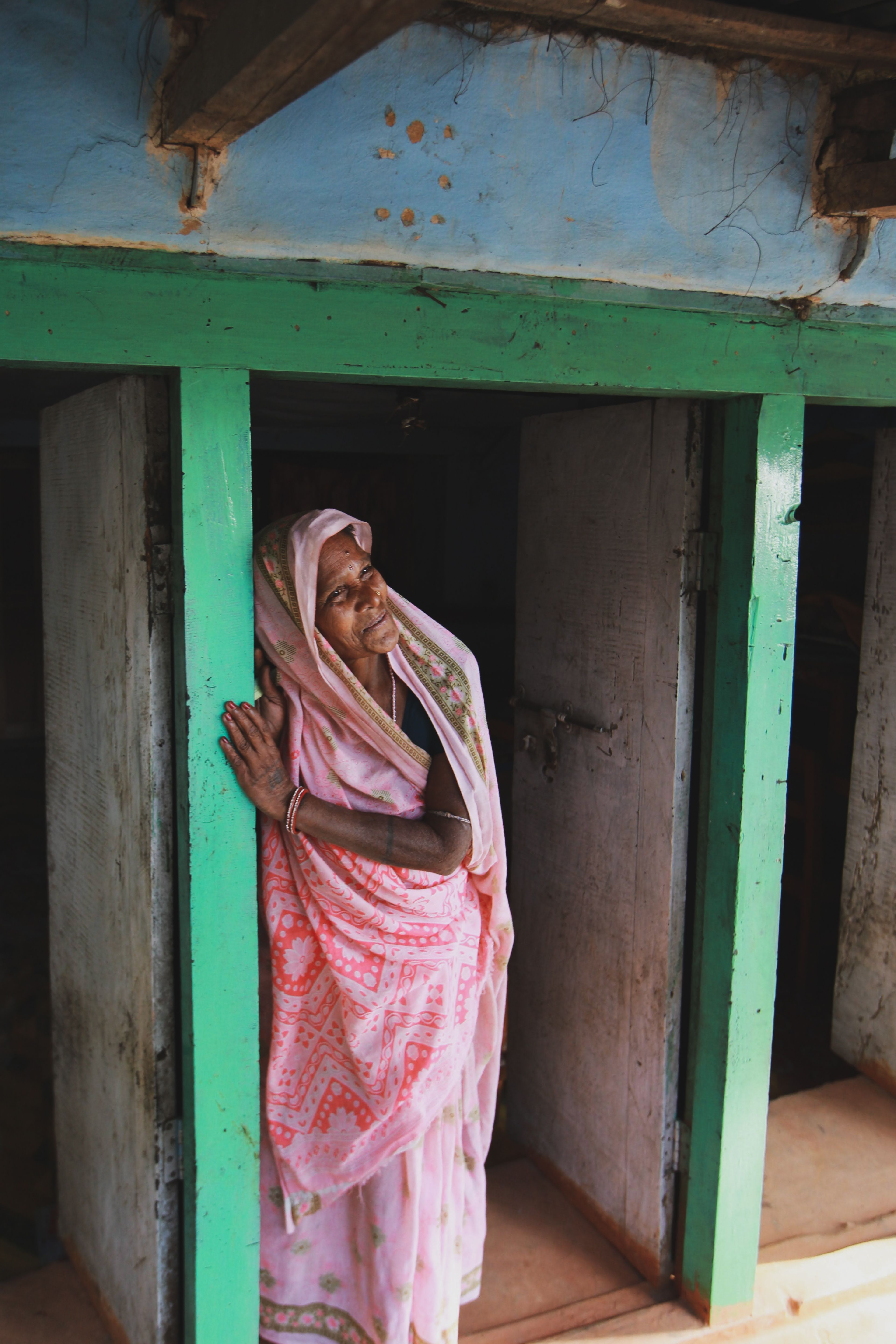 An elderly woman in traditional attire stands thoughtfully in a rustic doorway.