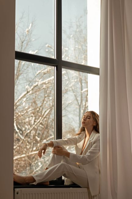Young woman in a white outfit sitting on a window sill with a snowy landscape outside in Almaty, Kazakhstan.