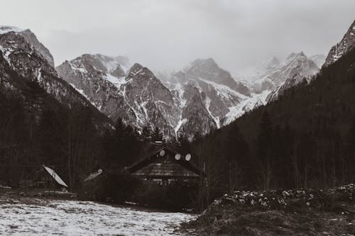 A Grayscale of a Cabin with Snow Covered Mountains on the Background