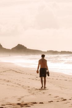 A shirtless man walks along a serene beach with cliffs in the background, capturing a peaceful coastal scene.