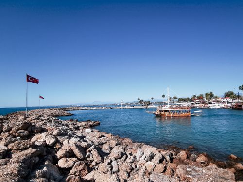 Free Picturesque view of a harbor with Turkish flags, stony shore, and a wooden boat sailing. Stock Photo