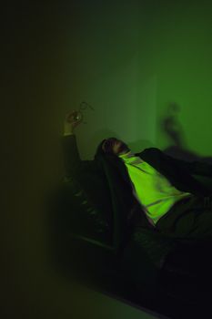 Man in a blazer reclines on a sofa holding eyeglasses, under dramatic green lighting.