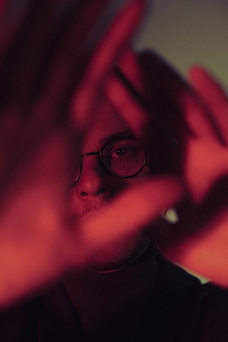 Young Man Looking Through Hole Made Of Hands In Red Light