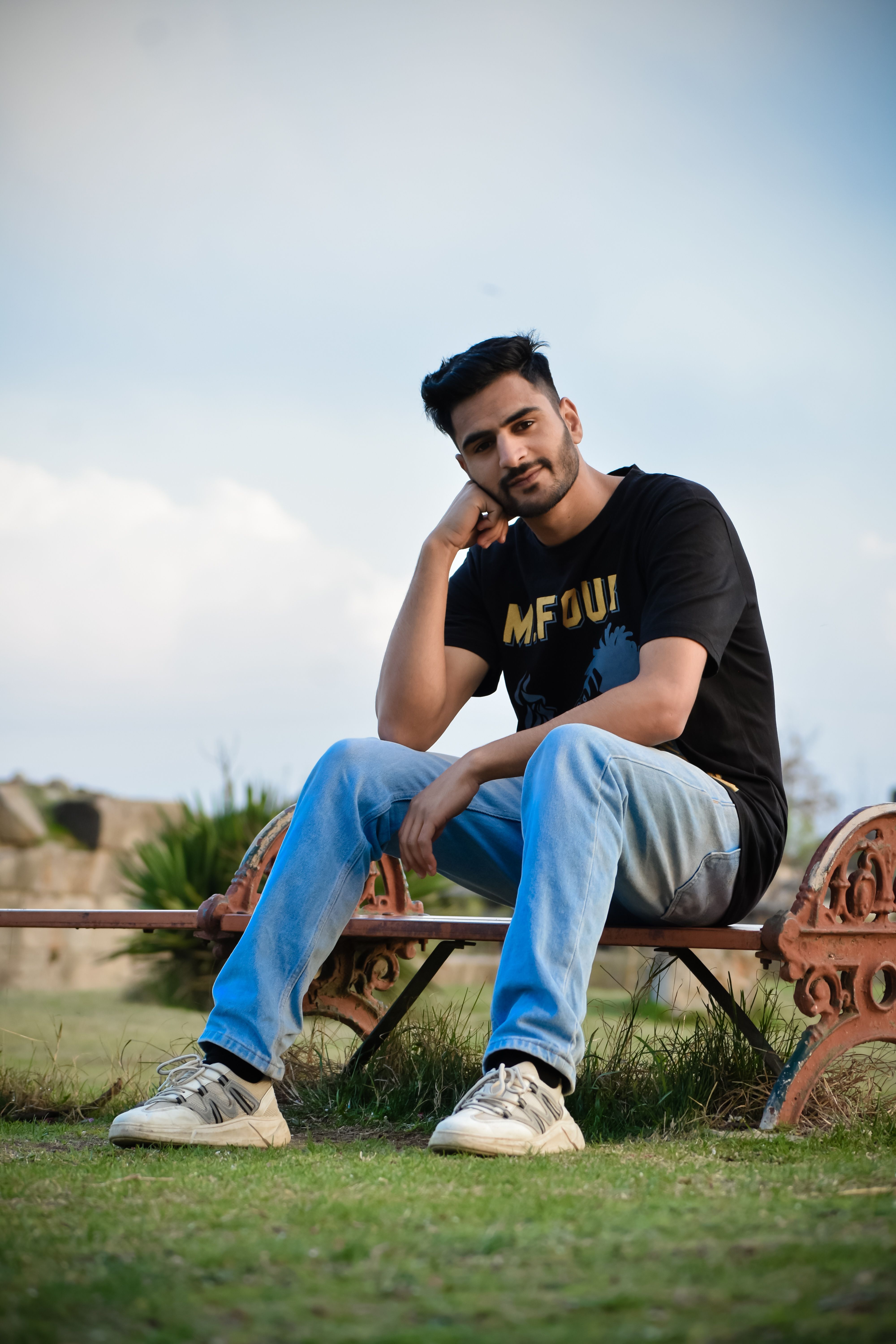 Casual portrait of a man in a black t-shirt and jeans sitting on a wooden bench outdoors.