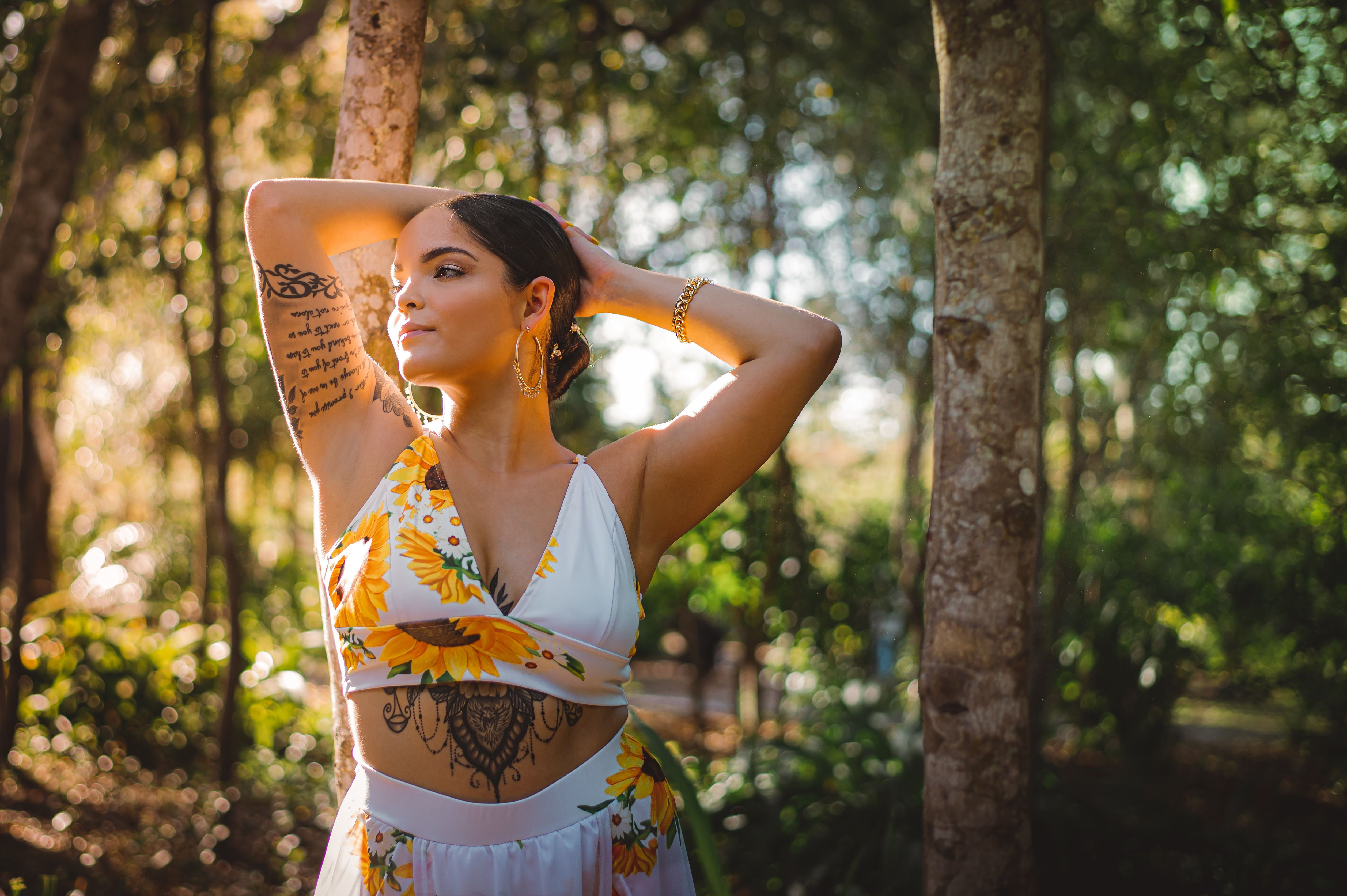 A woman with tattoos poses in a sunlit garden wearing a sunflower-themed outfit.