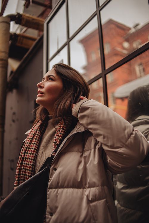 Free Stylish young woman looking up in city, wearing a jacket and scarf, urban scene. Stock Photo