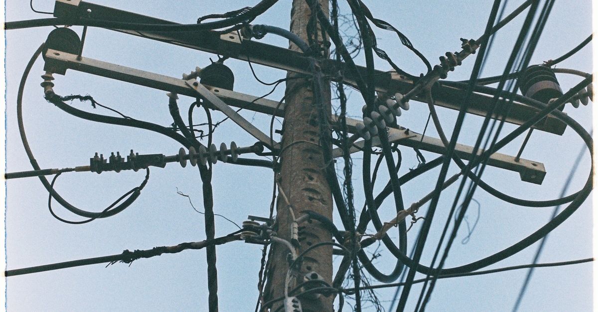 Photo by Nghia .8pm Complex web of utility wires on a wooden pole against a clear blue sky.