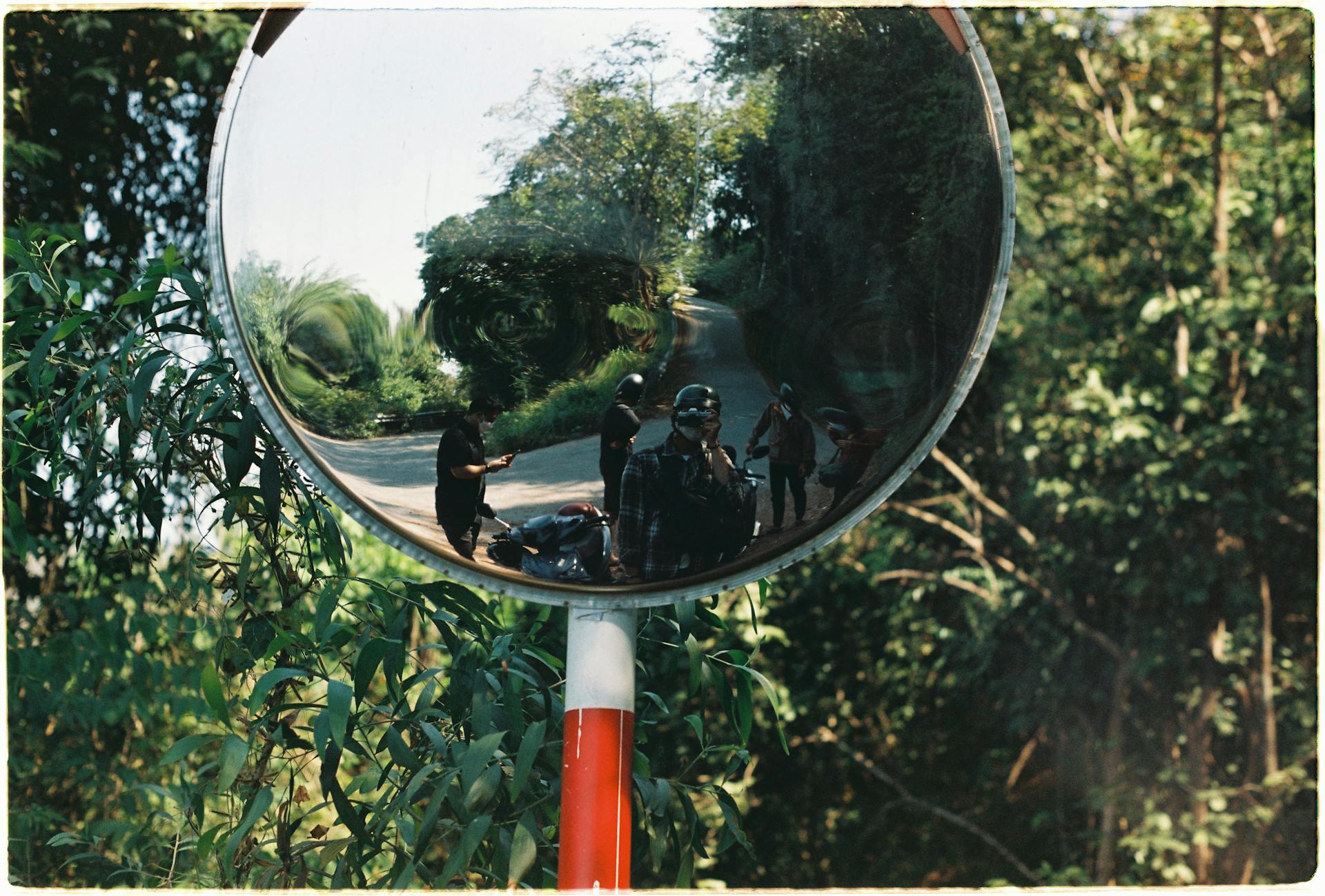 Group of bikers reflected in a convex mirror on a forest road, capturing nature and travel vibe.