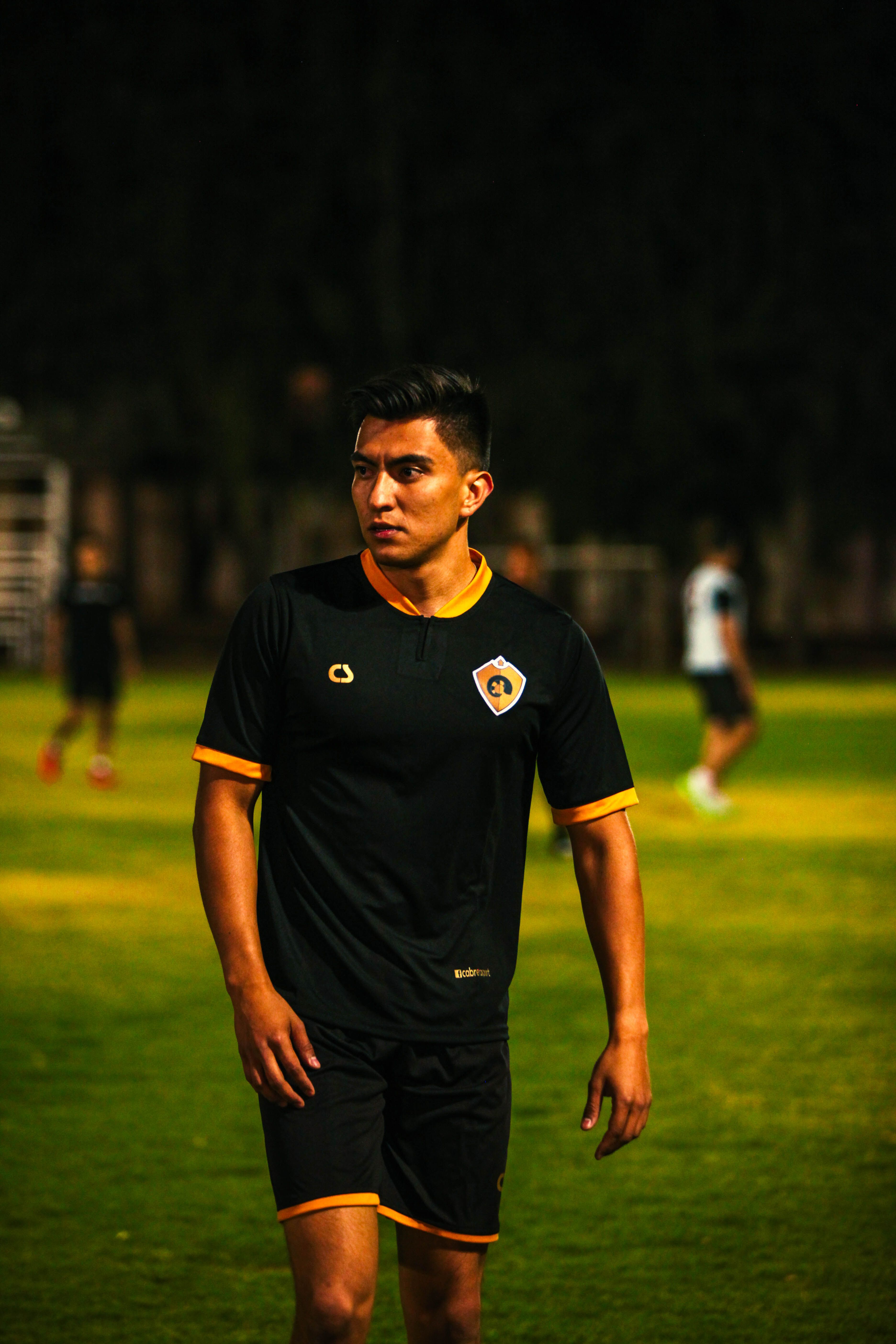 Focused young male soccer player in black jersey walking on field during night practice.