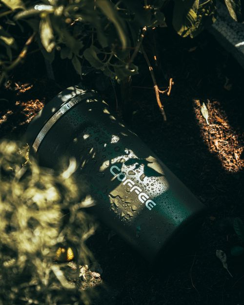 Free A coffee thermos with water droplets, placed outdoors amidst greenery, capturing the essence of freshness. Stock Photo