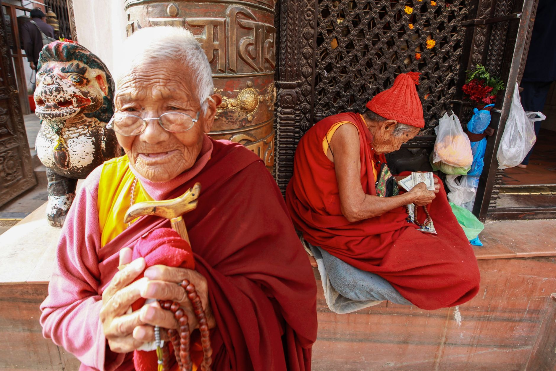 Two elderly Buddhist monks in traditional robes sit outside a temple, one holding prayer beads.