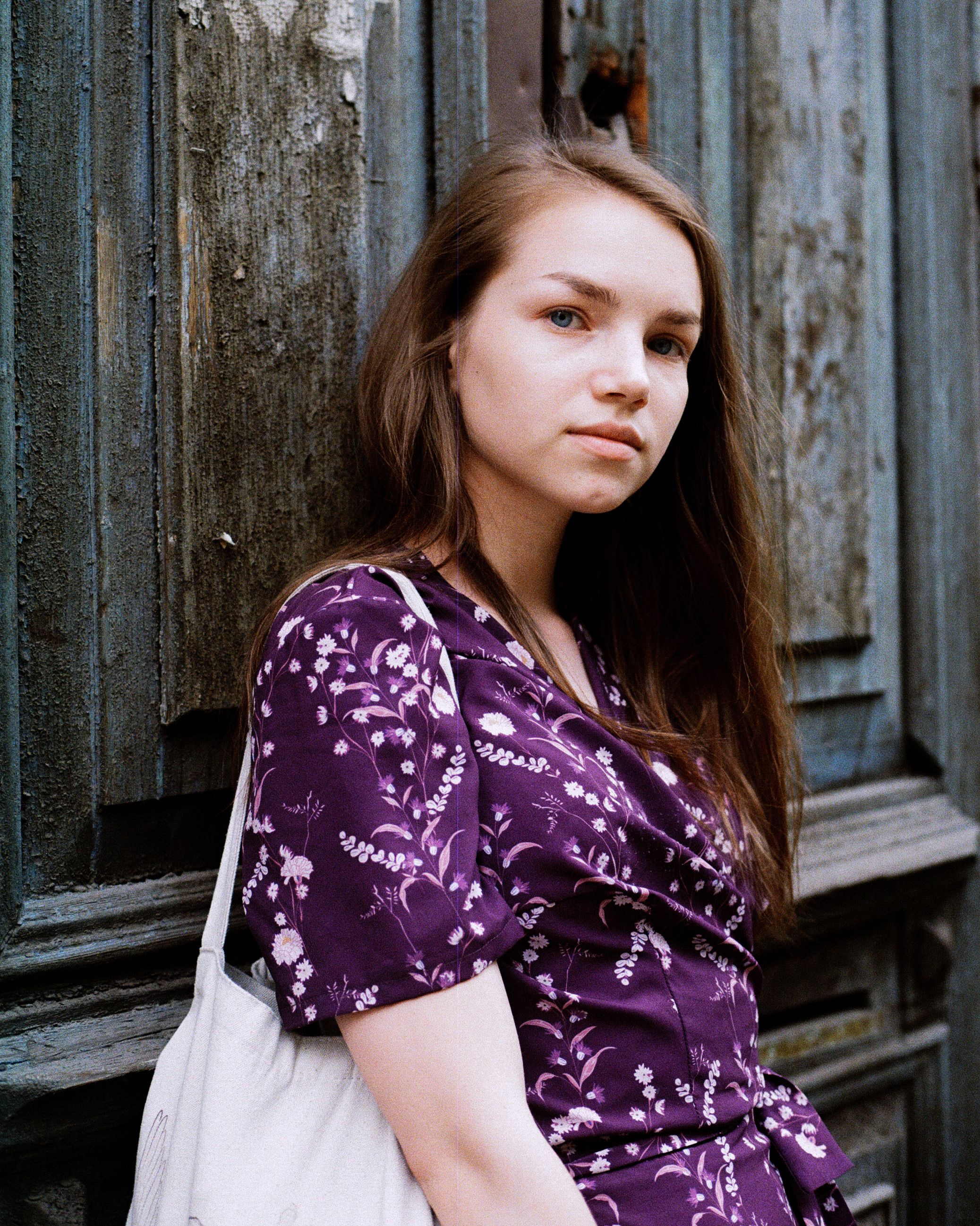 Portrait of a young woman in a purple blouse leaning against a weathered wooden door.