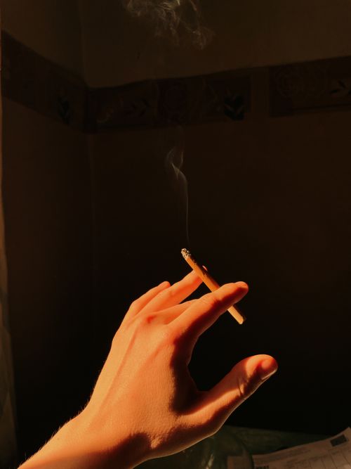 Free Close-up of a hand holding a cigarette with smoke rising, in warm lighting. Stock Photo