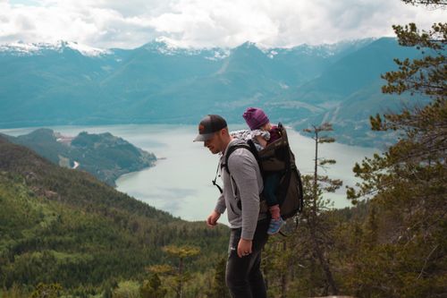 Free A father carries his child on a mountain hike with breathtaking views of a lake and mountains. Stock Photo