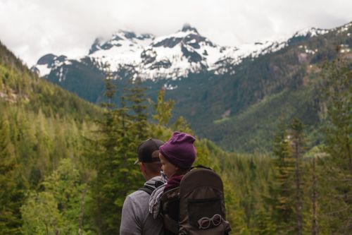 Free A father carrying his child while hiking in a scenic snowy mountain landscape with lush trees. Stock Photo