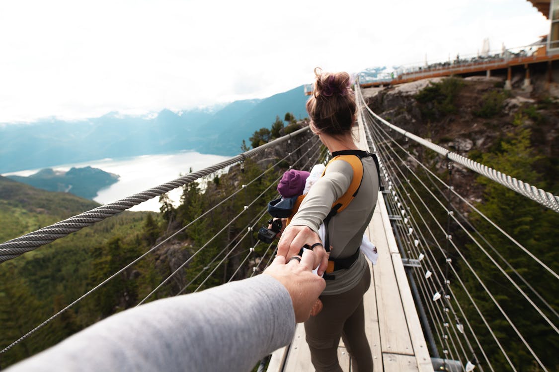Free Woman With Yellow Backpack Standing on Hanging Bridge With Trees Stock Photo