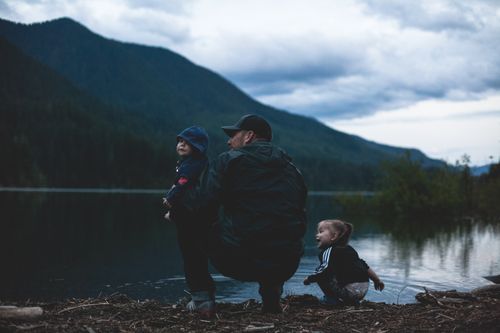 Free Father and children enjoy a serene moment by a lakeside with mountains. Stock Photo