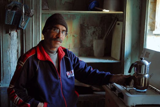 An adult male boils water on a gas stove in a rustic kitchen setting.