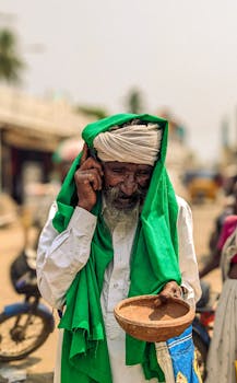 An elderly man in traditional clothing holding a phone and bowl, standing outdoors smiling.