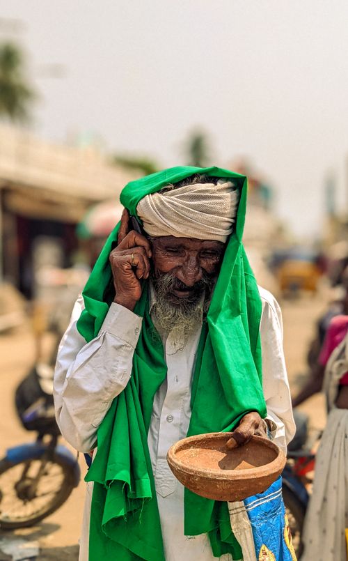 Free An elderly man in traditional clothing holding a phone and bowl, standing outdoors smiling. Stock Photo