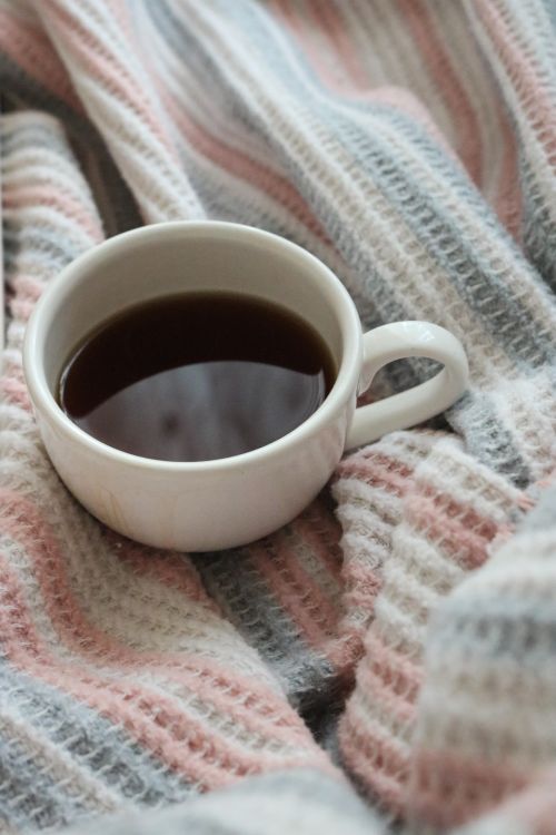 Free A warm ceramic cup of coffee nestled on a textured, pastel-striped blanket. Stock Photo