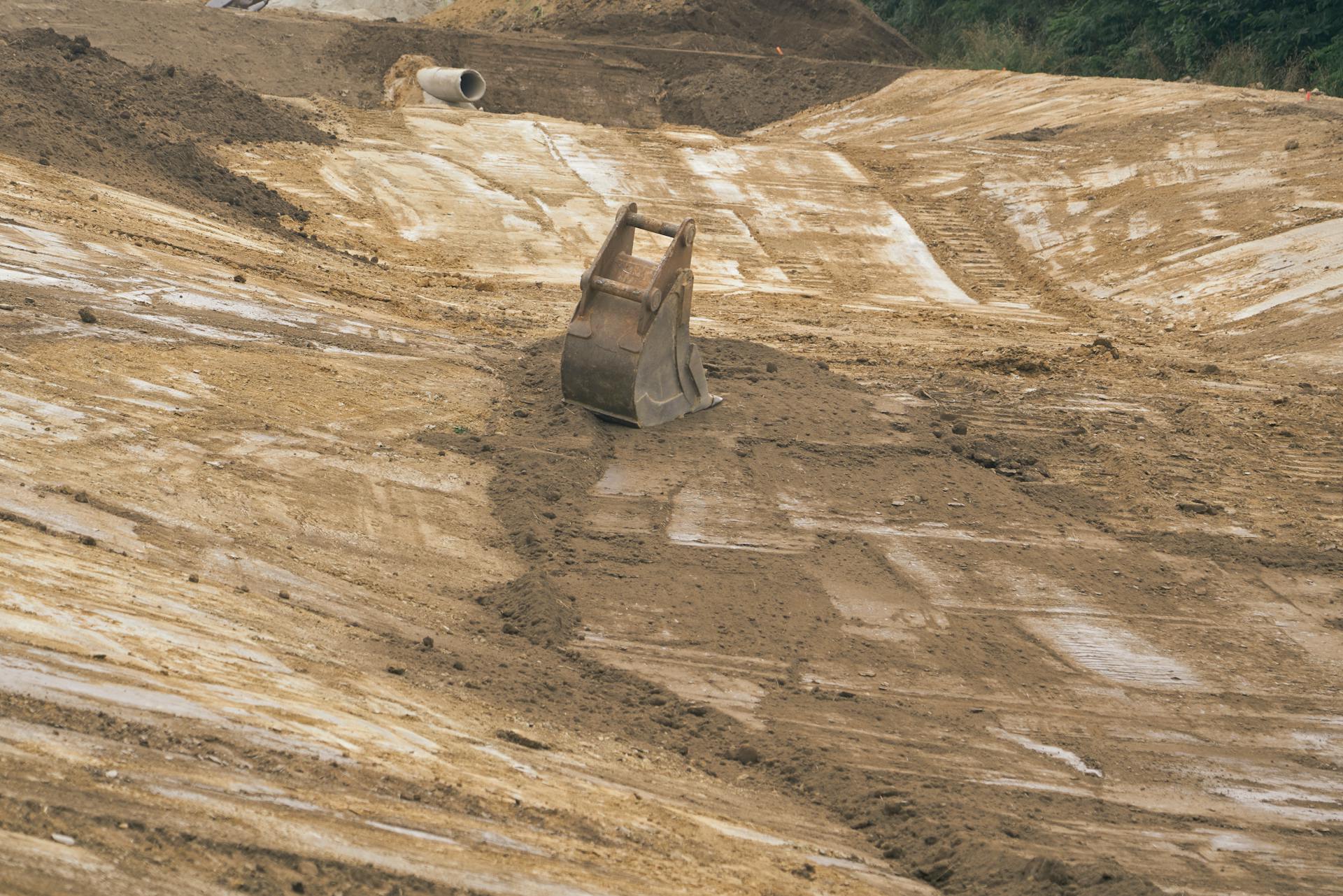 Excavator bucket at an outdoor construction site with dirt and equipment.