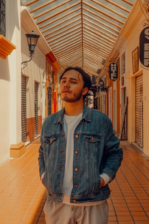Free A fashionable man in a denim jacket poses in a sunny outdoor corridor. Stock Photo