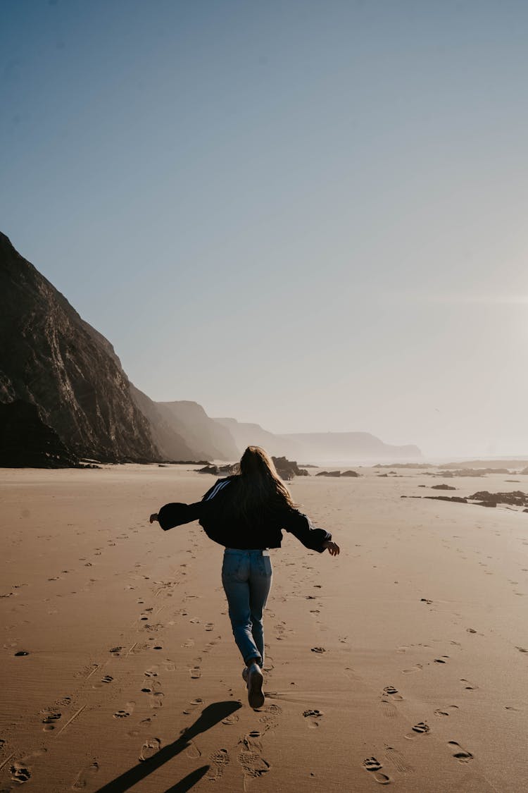 Woman In Black Jacket And Blue Denim Jeans Running On Brown Sand 