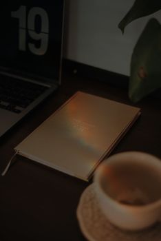 A warm, moody photo of a notebook, coffee cup, and laptop in a dimly lit workspace.