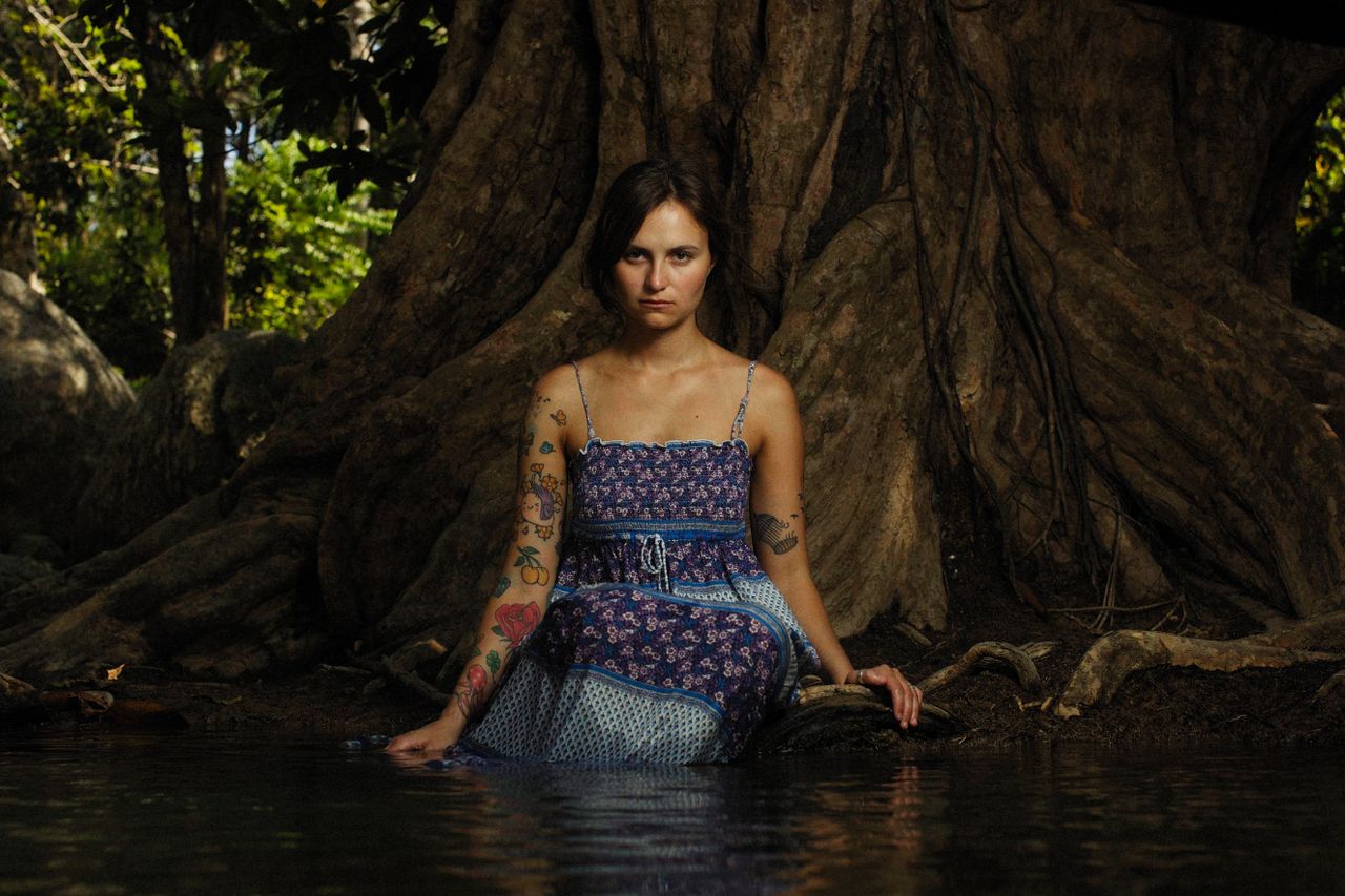 a serious woman with tattoos sits by a tree in nature partially submer