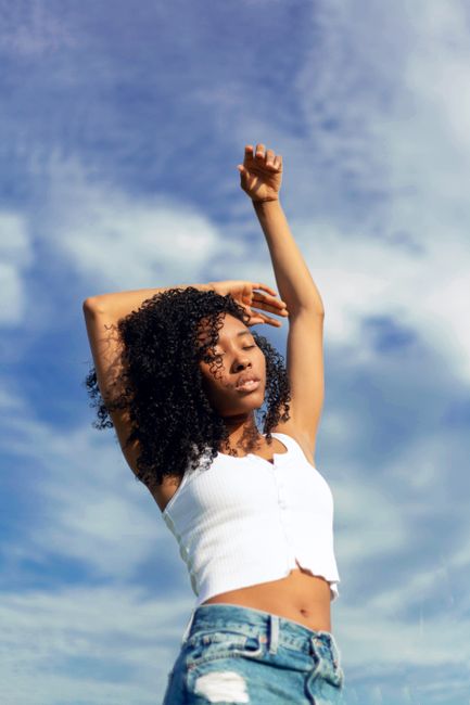An adult woman with curly hair poses confidently against a vibrant blue sky, embracing a carefree summer vibe.