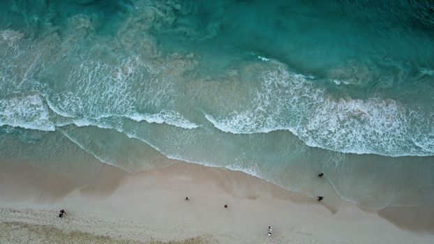 Drone shot capturing the serene waves, sandy beach, and scattered people from above.