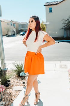 Stylish woman with long red hair in an orange skirt standing in an urban neighborhood on a sunny day.