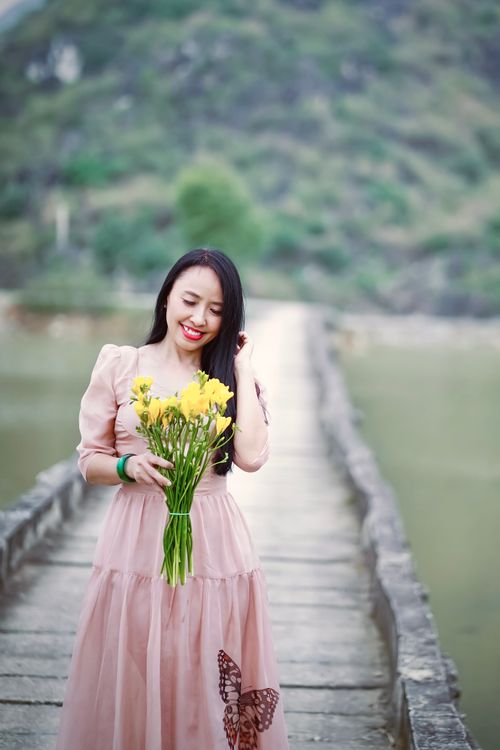 Free A joyful woman in a pink dress holds yellow flowers while standing on a picturesque bridge with a scenic background. Stock Photo