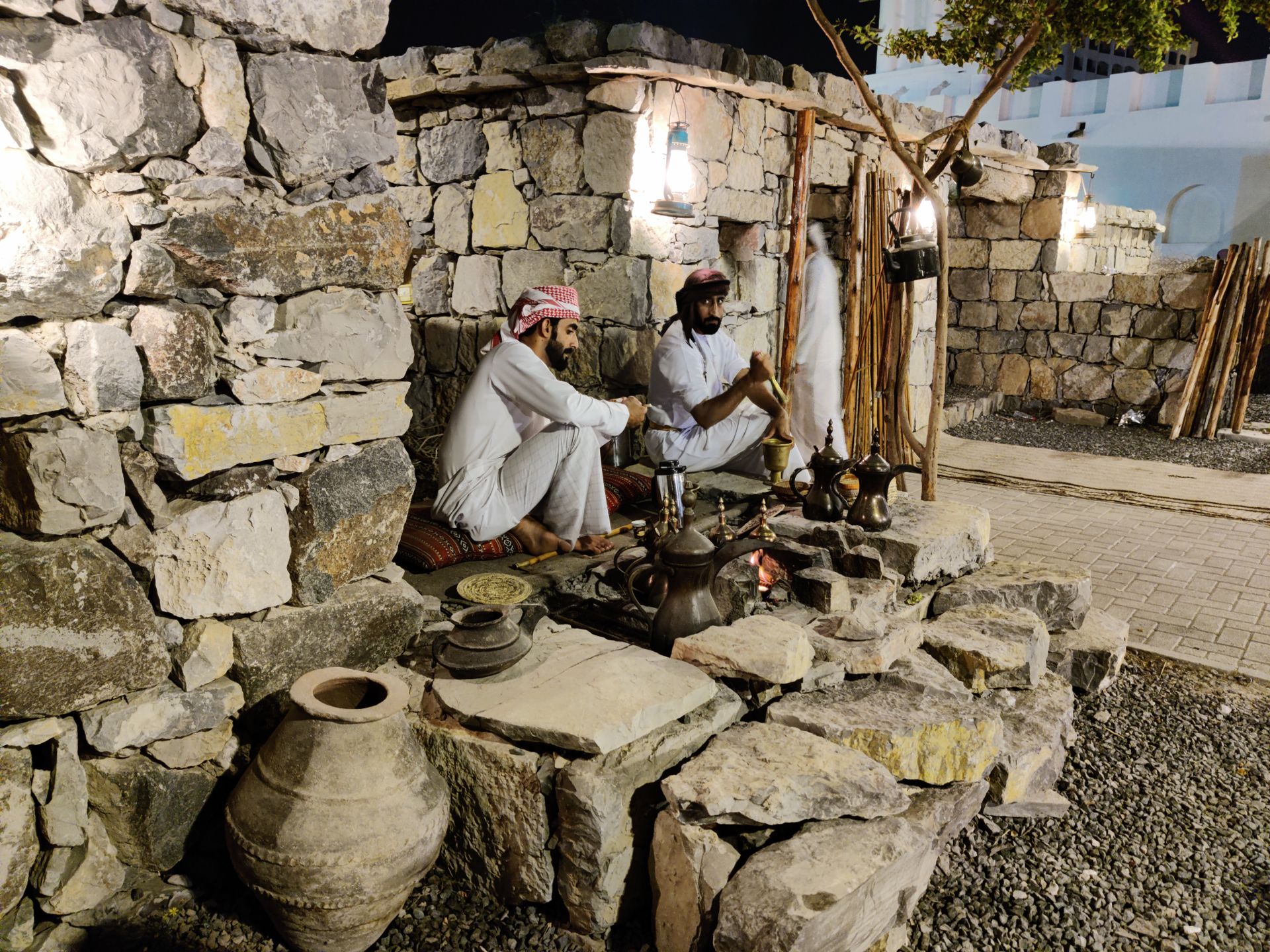 Two Arab men sit by a fire in a rustic setting, preparing traditional coffee pots at night.
