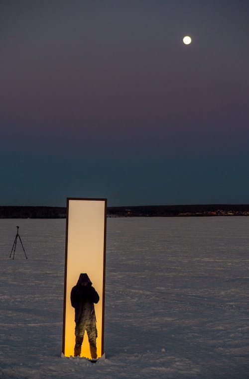 镜子中映出男人和金色天空的轮廓，镜子矗立在结冰的海冰上，背景是粉紫色的天空和月亮