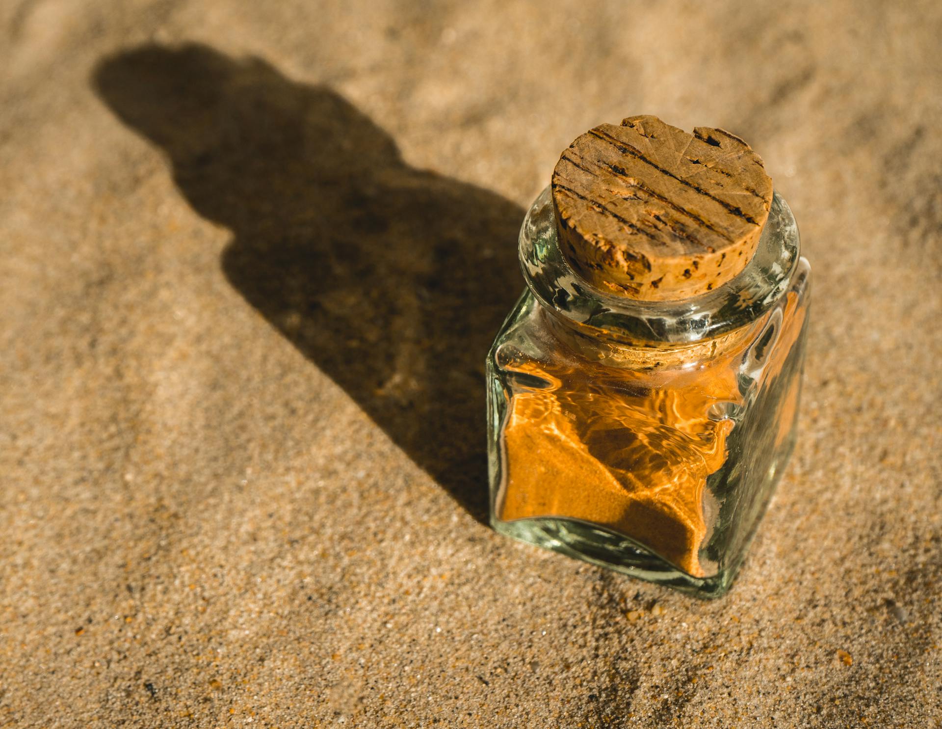 A glass bottle sealed with a cork, containing sand, casting a shadow on a sunlit surface.