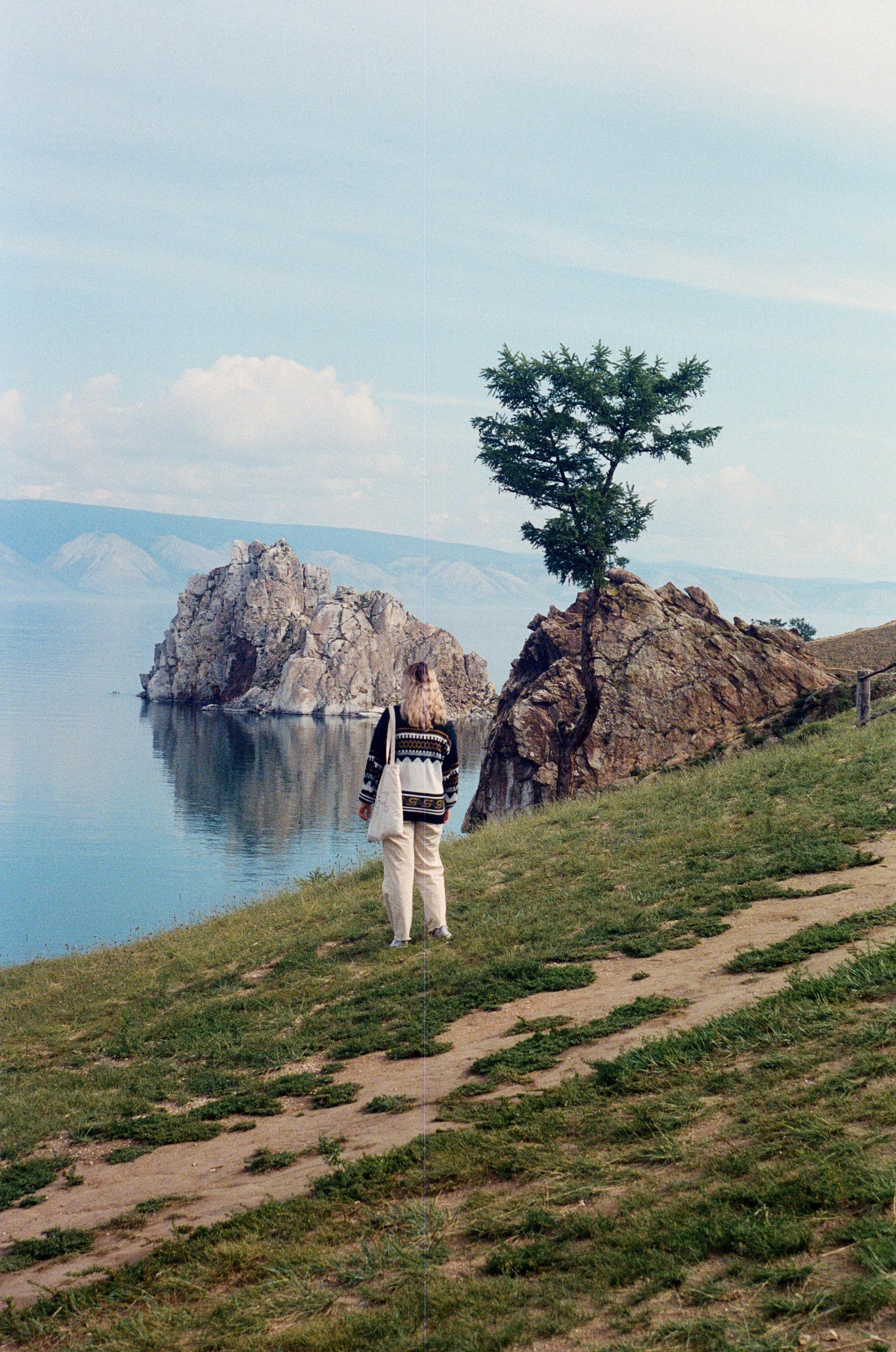 Woman standing on grassy hill, overlooking serene lake and rocky formations under a blue sky.