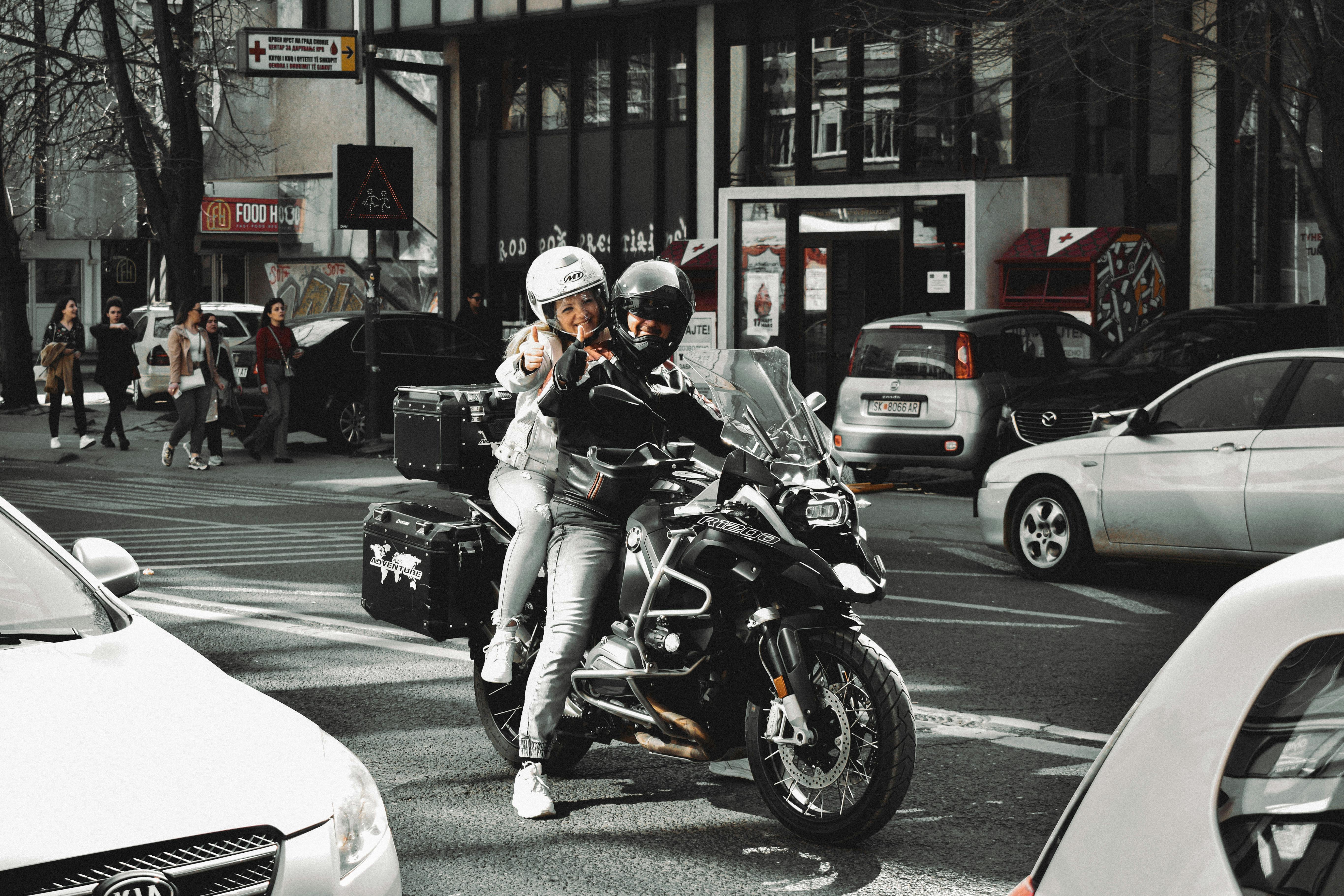 A couple on a motorcycle gives a thumbs up while riding through city traffic.